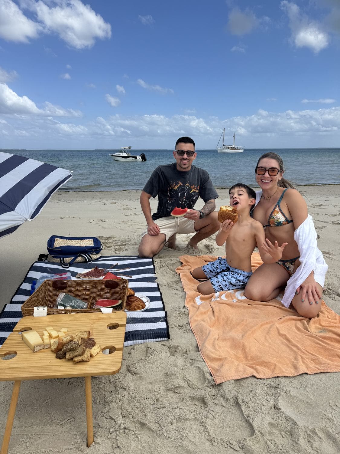 A family picnic on the sand with a striped umbrella, wicker basket and pastries
