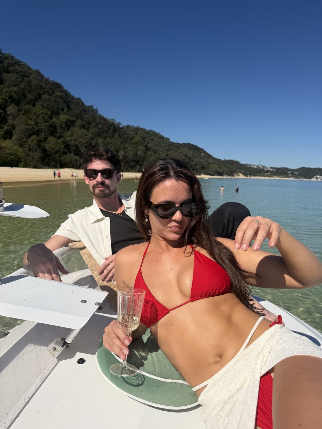 A couple relaxing in the cockpit with champagne, the beach and clear water behind